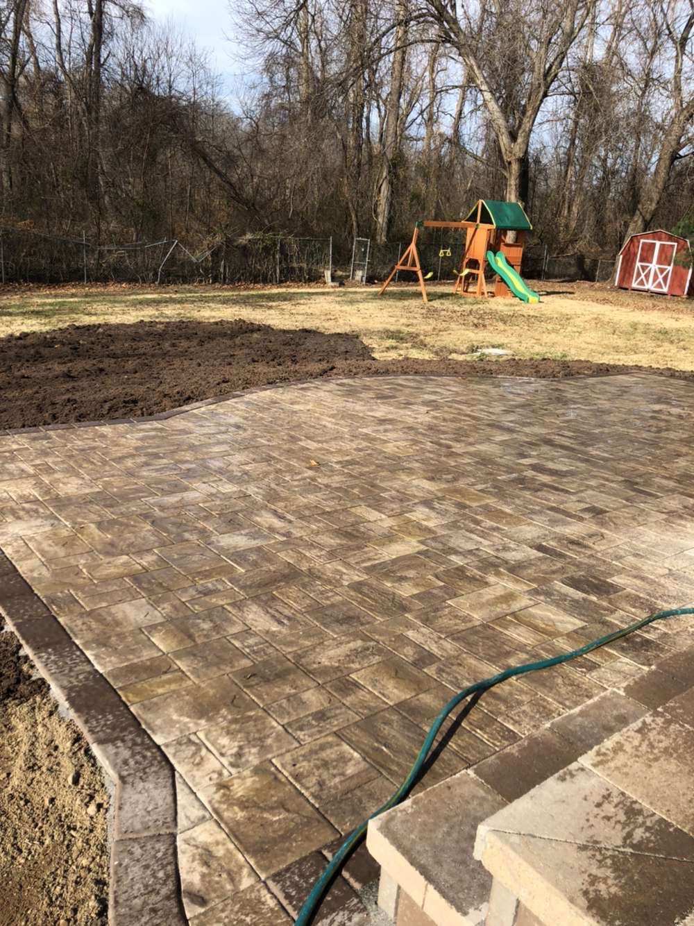 Backyard patio with a green hose, playset, leafless trees, and a small red shed in the background