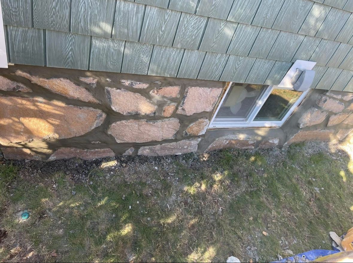 Exterior wall with stone foundation, gray shingles, and a small window beside a patchy grass yard.