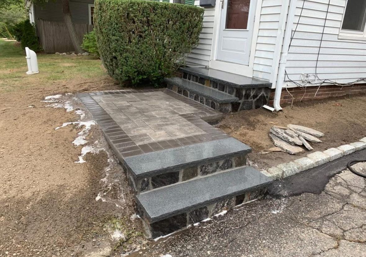 Small concrete steps leading to a white house entrance beside a bush and cracked driveway.