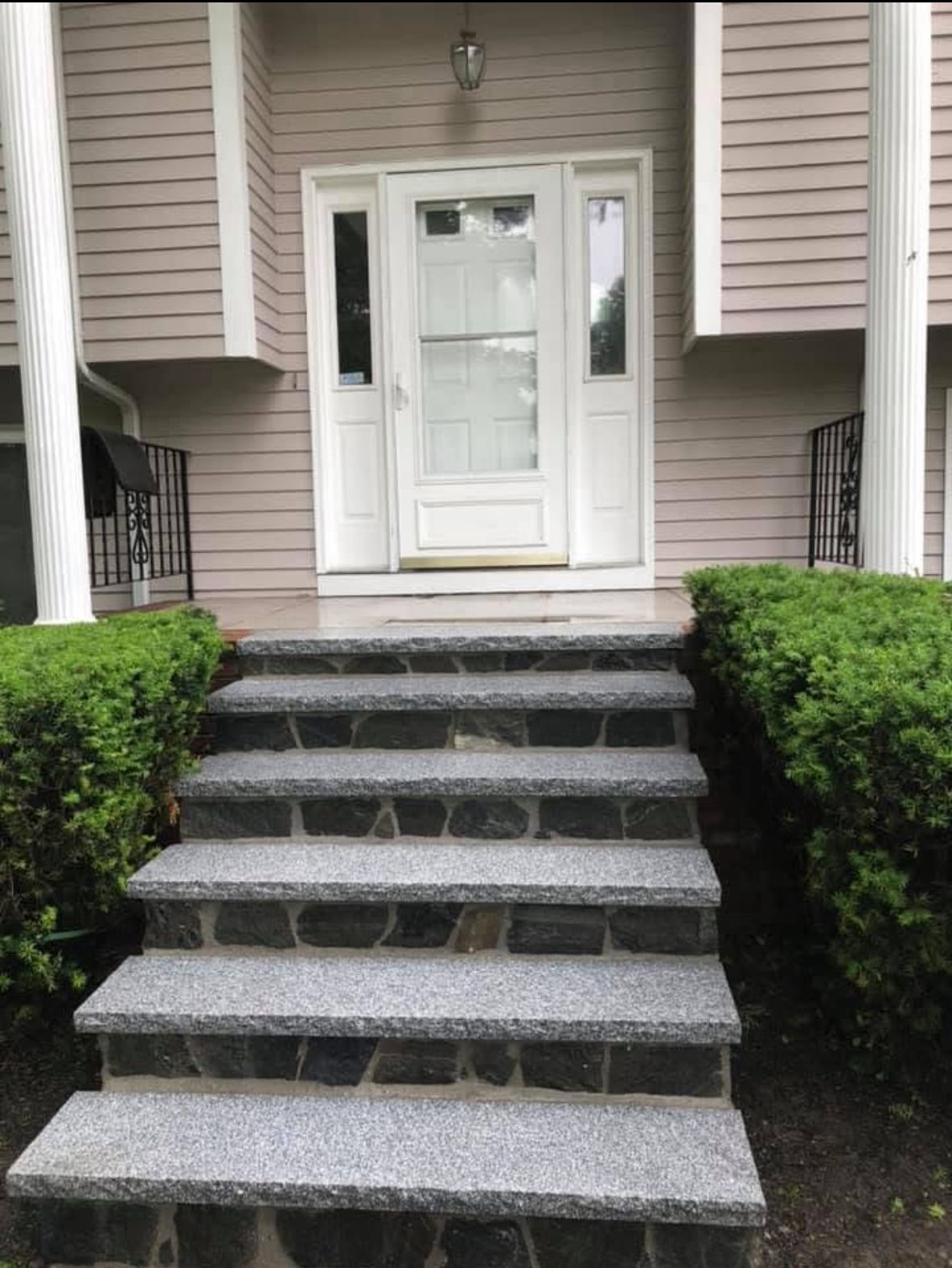 Front entrance with white door, gray stone steps, and trimmed green bushes on both sides