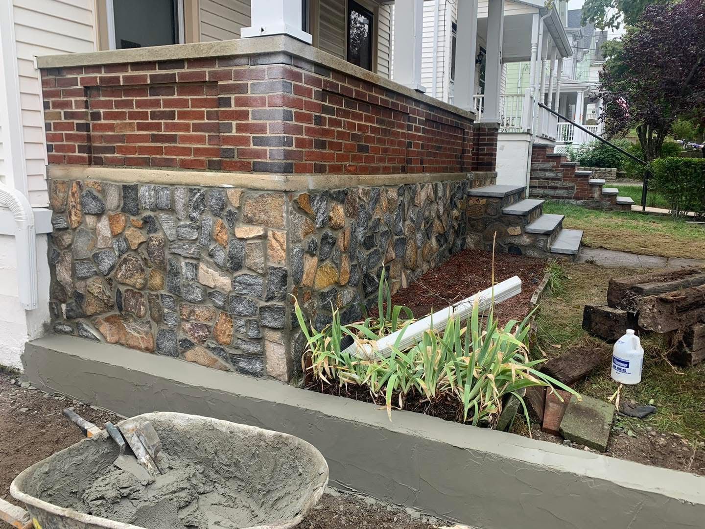 House corner with brick-and-stone foundation, concrete walkway, steps, and a small planted garden.