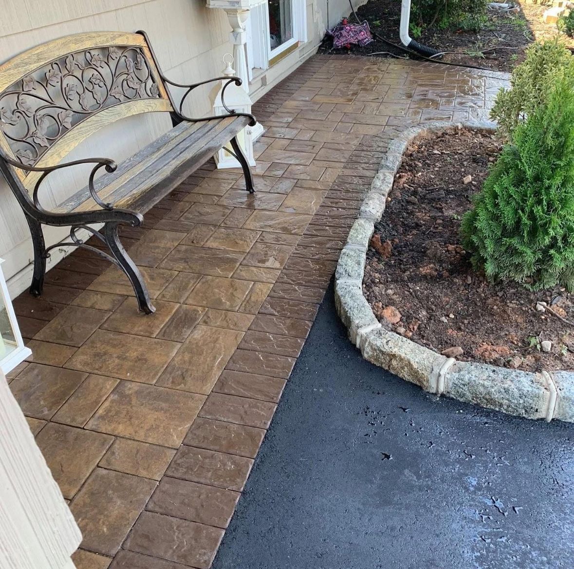 Curved brick patio beside a bench and landscaped bed, meeting a black asphalt walkway.