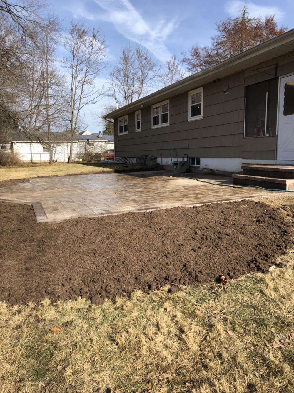 Backyard with a freshly graded soil bed beside a house and concrete patio, under a blue sky