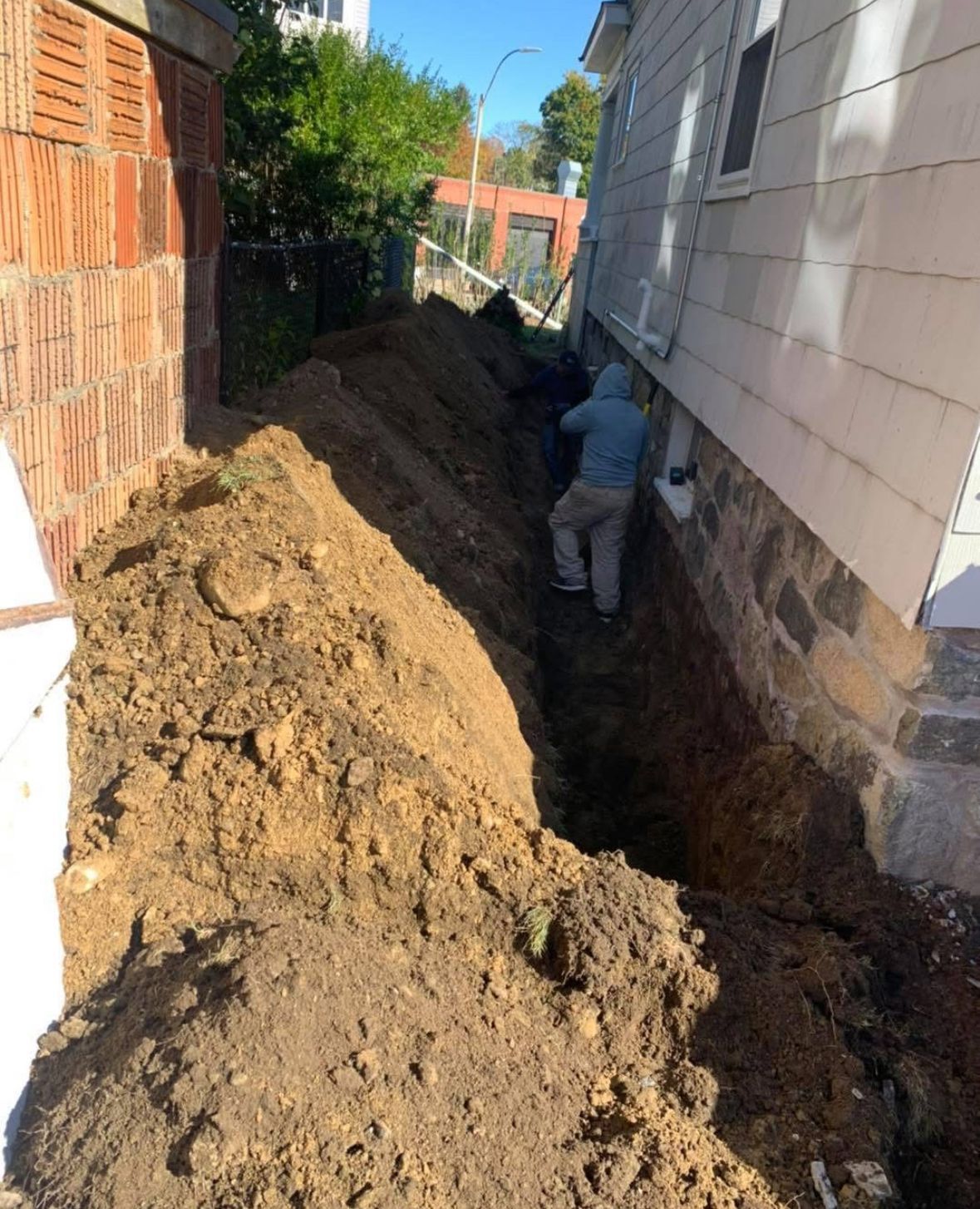 Worker digging a trench beside a house, with a large dirt pile along the narrow path