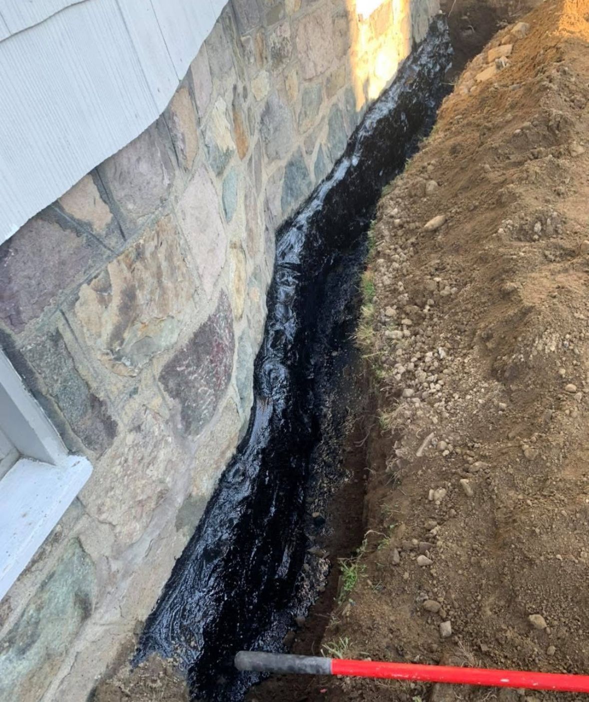 Black waterproofing along a stone foundation beside a dirt trench, with a red-handled tool in the foreground