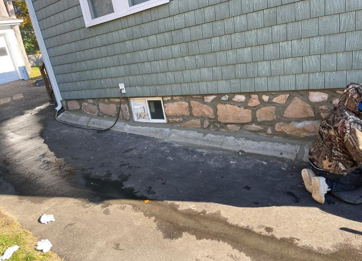 Basement wall with exposed stone foundation, dark paved strip, and a window well beside a driveway.