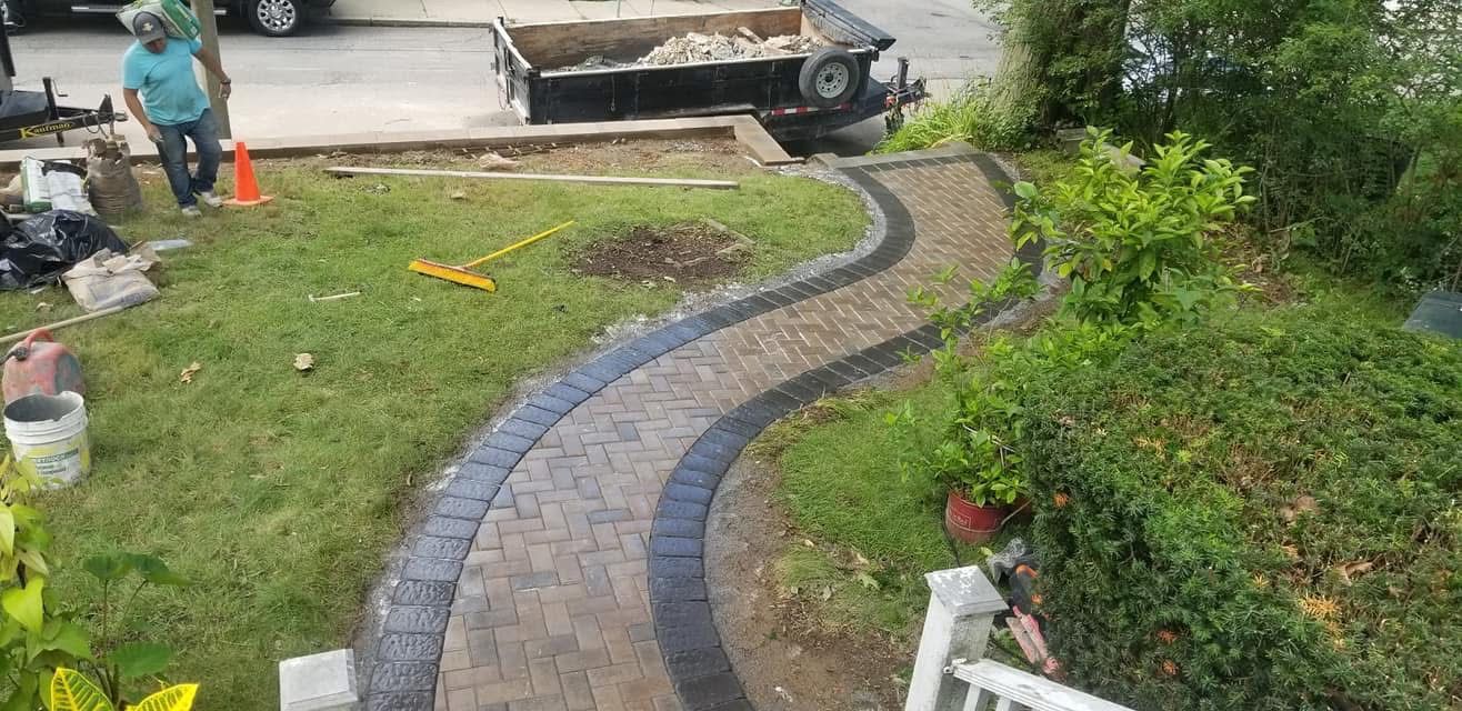 Curved brick garden path bordered by grass and shrubs, with a person and wheelbarrow in the background