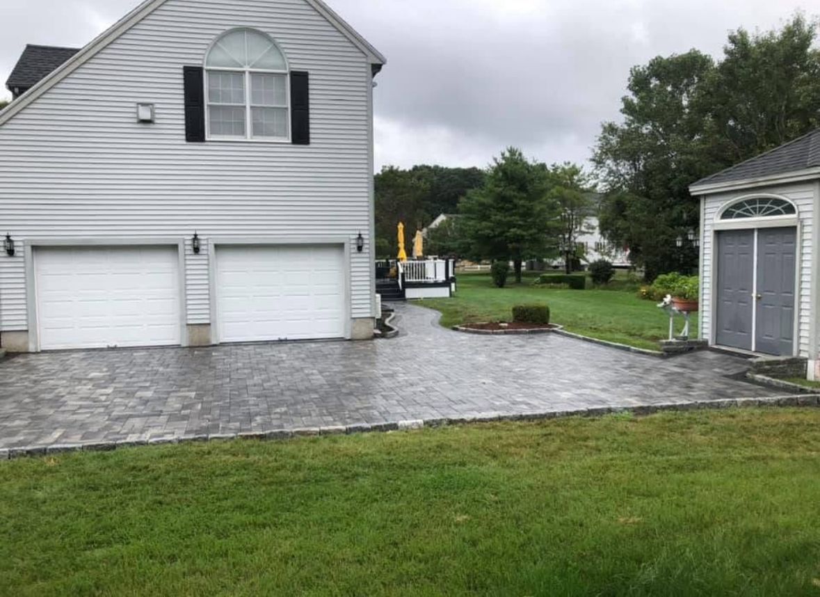 White house with two garage doors, stone driveway, and lawn after rain; small shed at right.