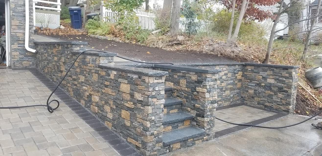 Stone outdoor wall and raised planter beside a paved walkway, with a white railing and steps nearby