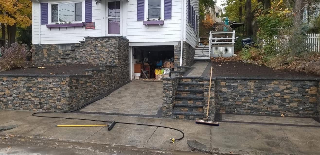 Stone retaining walls and stairs leading to a house entrance, with a concrete driveway in front.