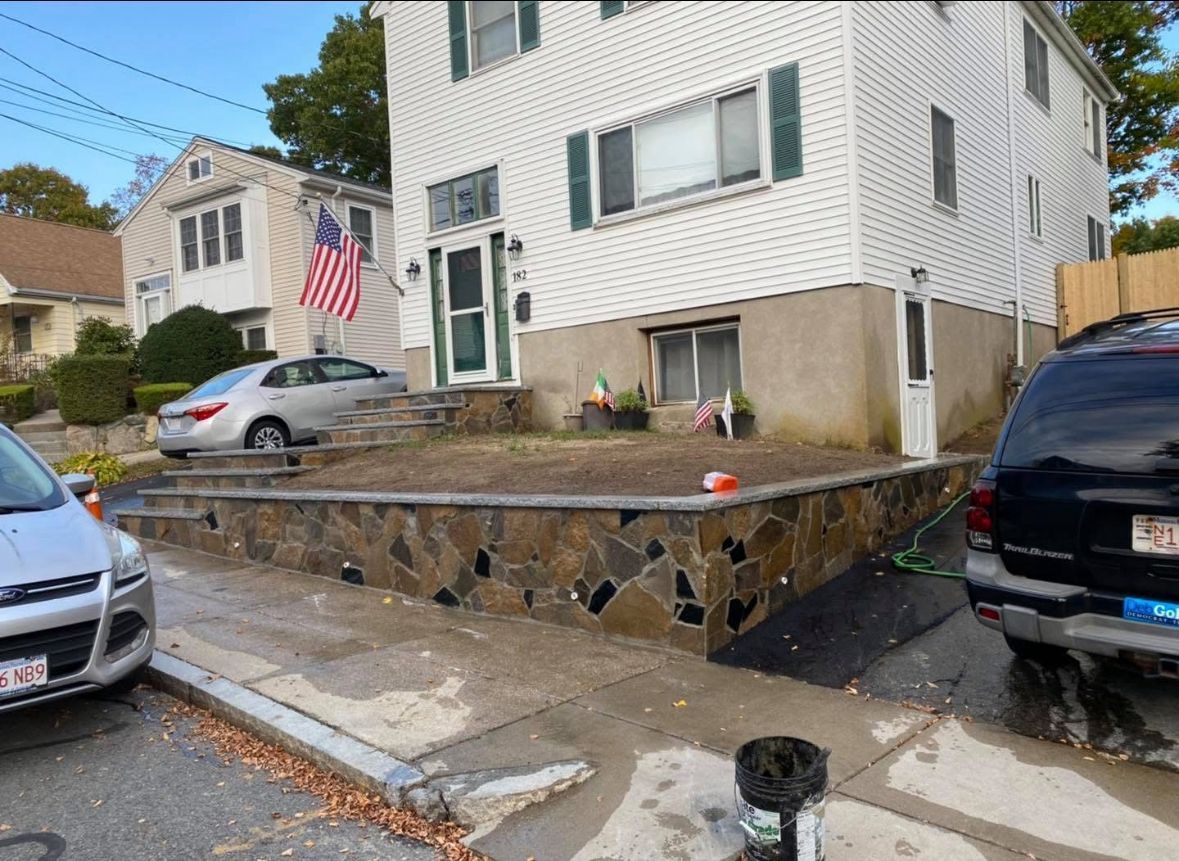 Residential corner lot with a white house, parked cars, and a stone retaining wall beside a sidewalk.