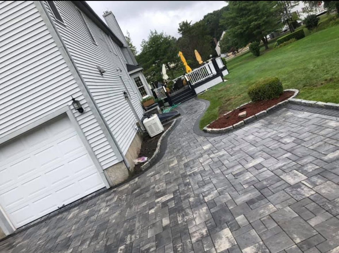 Tilted view of a house driveway with gray pavers, garage door, lawn, and patio furniture in the background