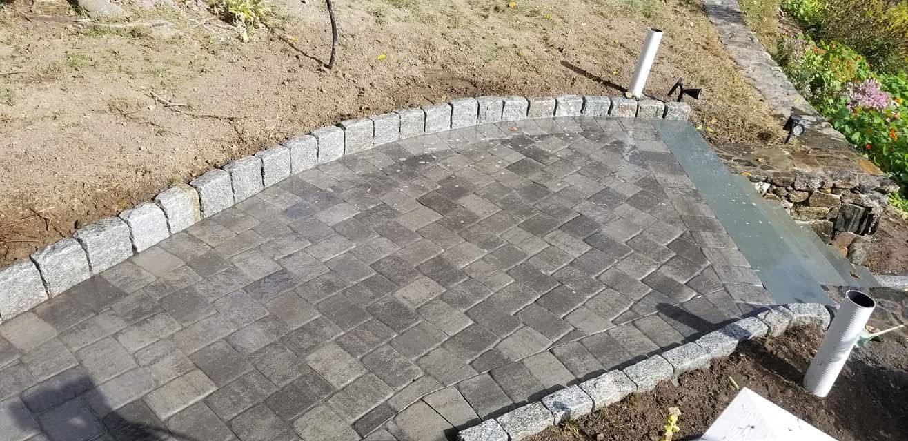 Curved paver patio bordered by stone edging on a sloped yard, with two white posts nearby.