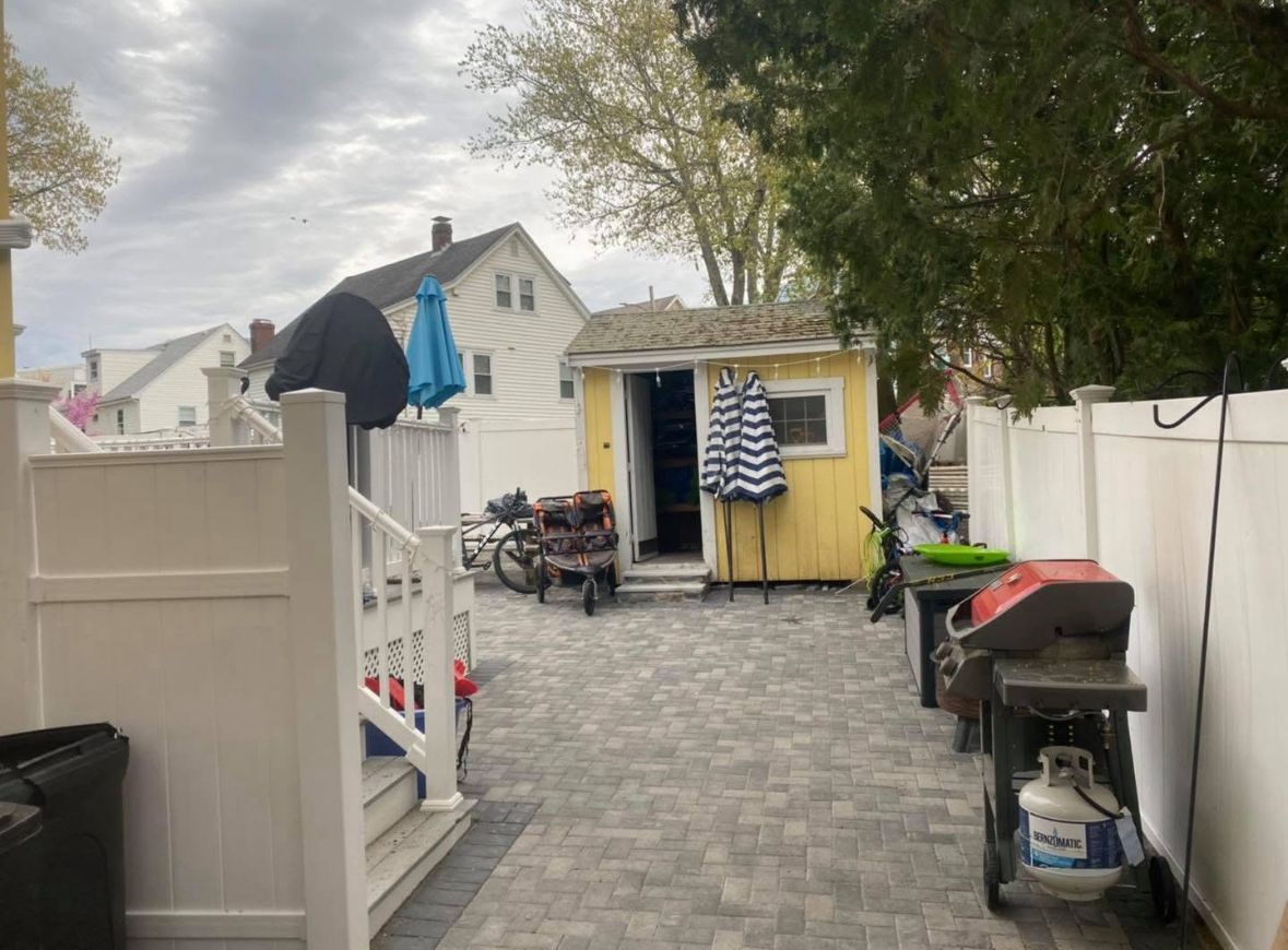 Backyard patio with pavers, white fence, yellow shed, and a motorcycle parked near hanging clothes.