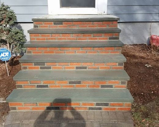 Brick front steps with gray concrete treads leading to a house entrance, bordered by mulch and a small sign.