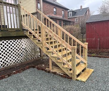 Wooden outdoor staircase with railing leading from a raised deck to a gravel yard beside a red shed.