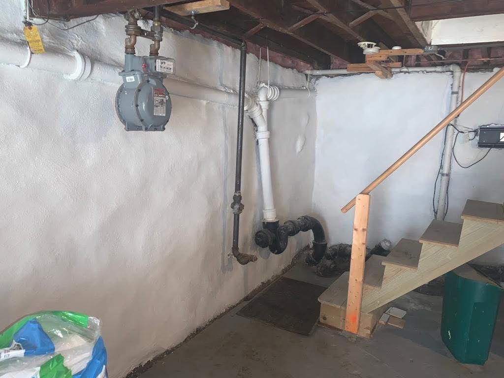 Basement utility area with exposed pipes, white walls, a staircase, and stored items on the floor.