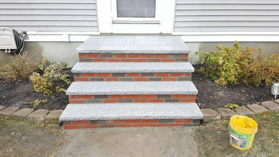 Front steps with gray concrete treads and red brick risers leading to a house entrance, bordered by shrubs.