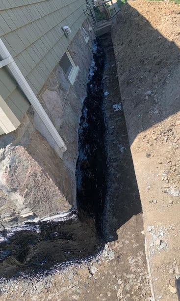 Narrow trench dug beside a house foundation, with exposed soil and a dark waterproofed corner.