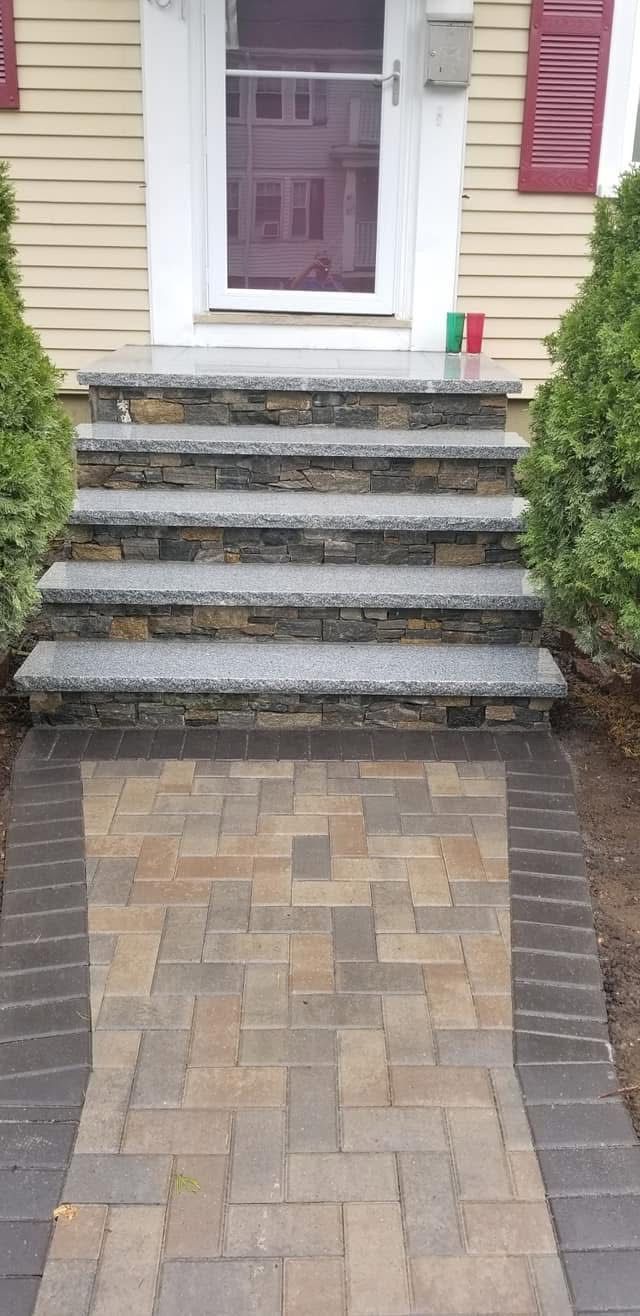 Front steps to a house with a brick walkway and white door, flanked by shrubs and maroon shutters