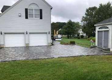 White house with three garage doors and a paved driveway beside a small yard and shed