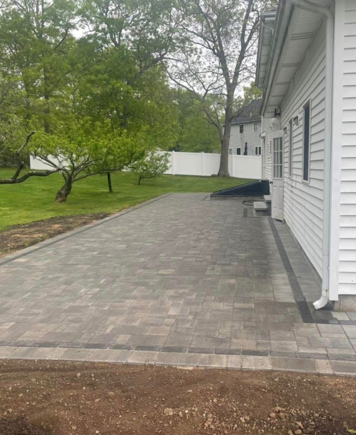 Paved patio beside a white house with a grassy yard and trees in the background