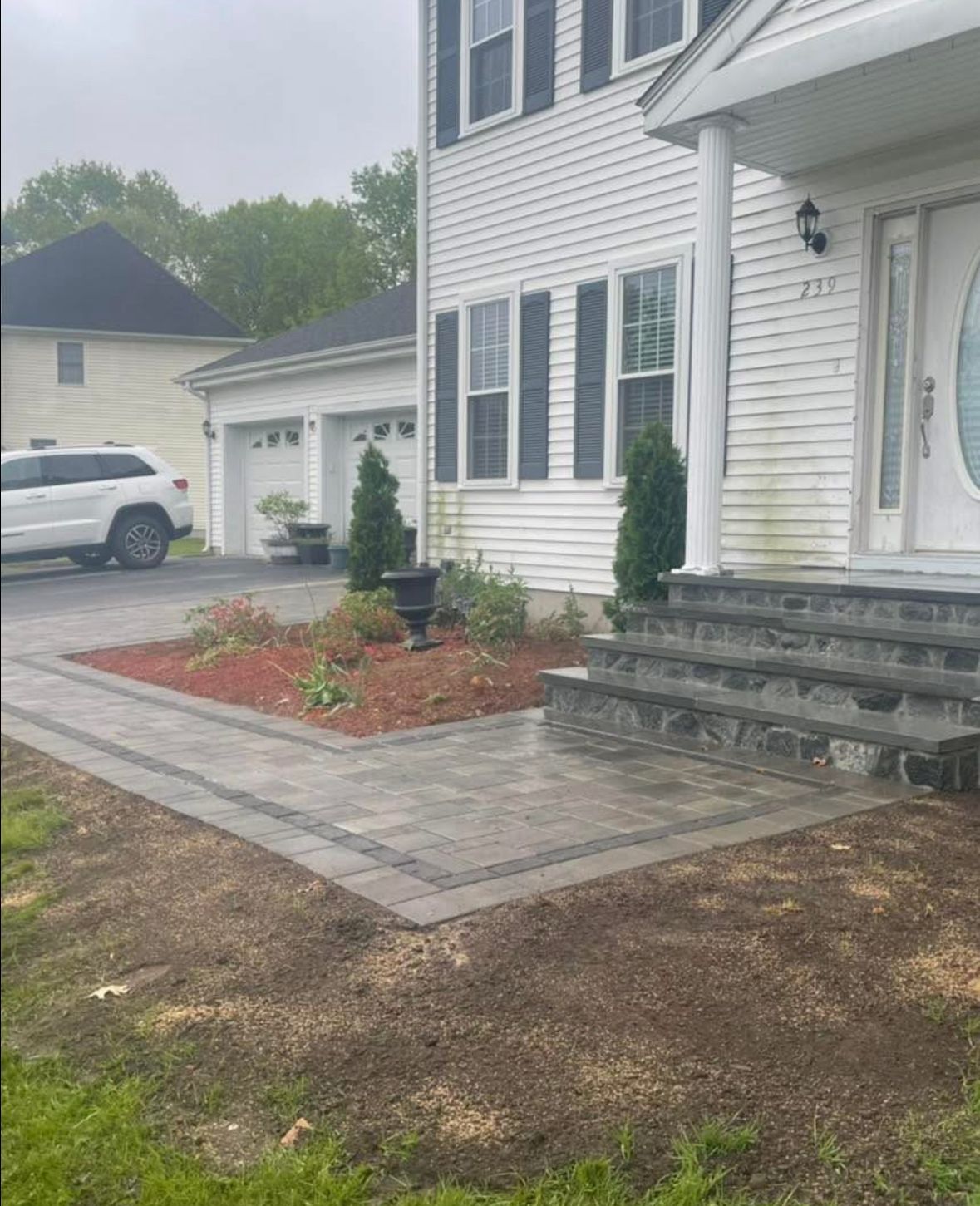 Front entry of a white house with gray steps, brick walkway, and landscaped flower bed beside a driveway.