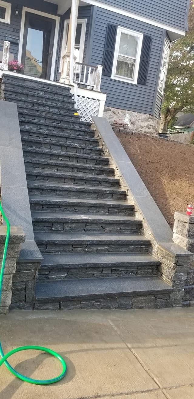 Front steps of a gray house with black railings, leading up from a sidewalk near a green hose.