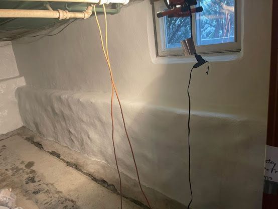 Basement wall with white spray foam insulation, exposed pipes, and a small window with hanging cords