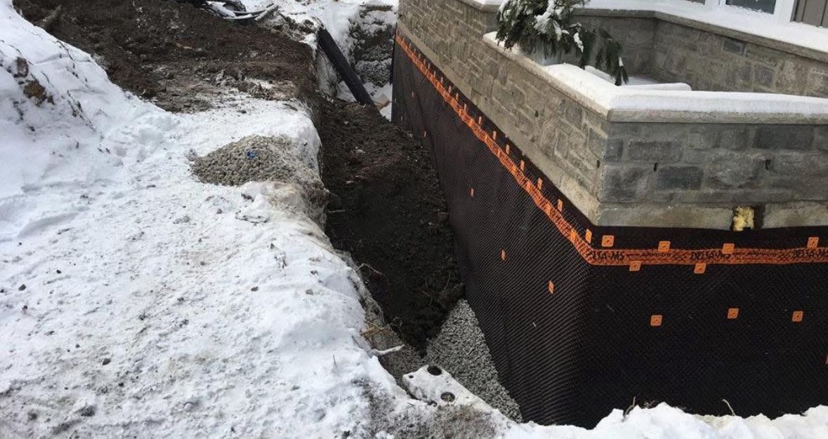 Snow-covered foundation with black waterproofing membrane and gravel trench beside a concrete structure