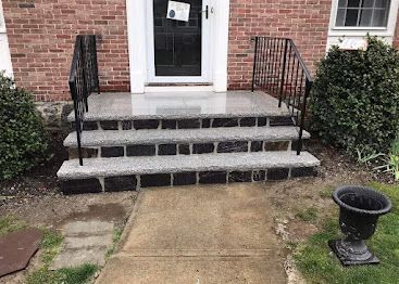 Brick house entrance with three concrete steps, black railings, and a front walkway.