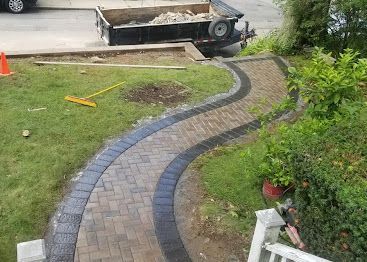 Curved gray brick walkway through a lawn, leading to a sidewalk with a wheelbarrow in the background.
