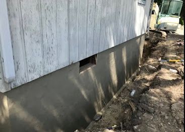 Side of a weathered building with a concrete foundation, dirt ground, and a truck in the background.