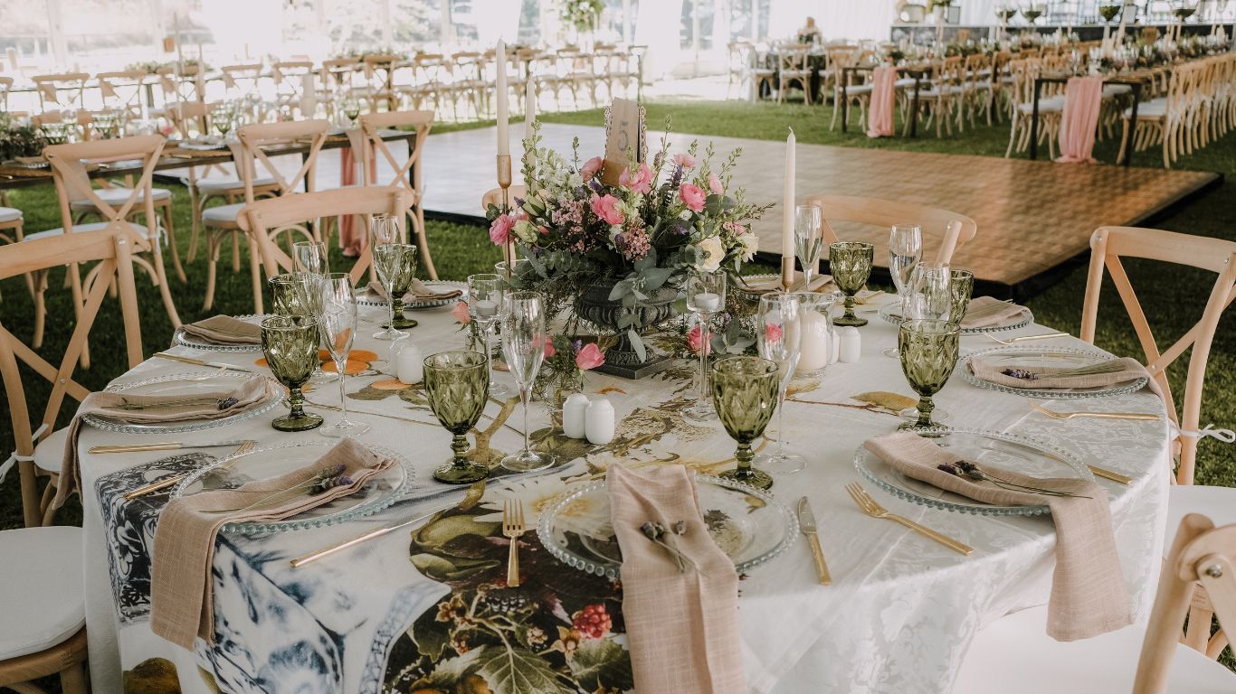Wedding reception table set with floral centerpiece, green glasses, and pink napkins; outdoor setting.