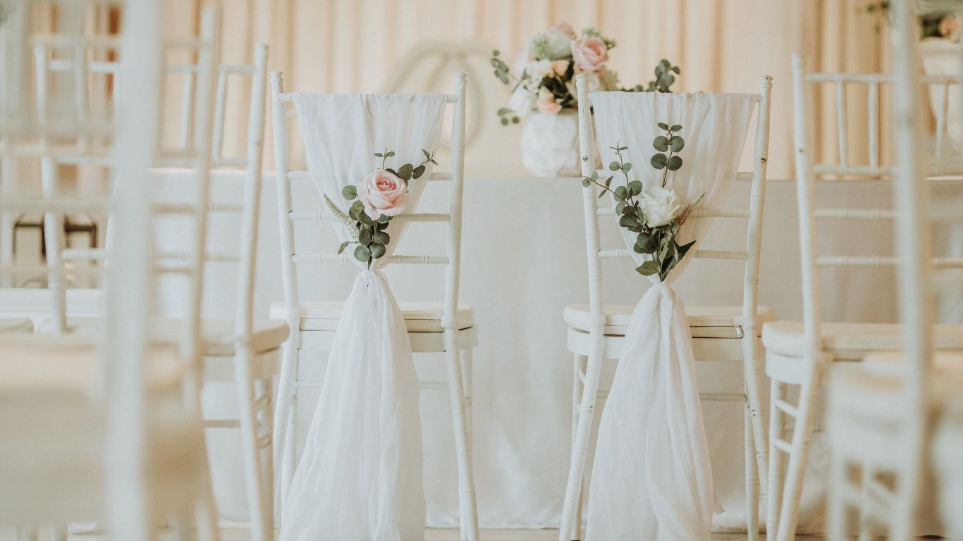 Two white chairs decorated with white fabric, flowers, and greenery, set for a wedding.