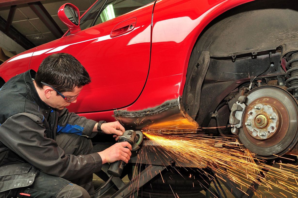 Mechanic using a grinder on a red car's side, sparks flying, wheel visible.