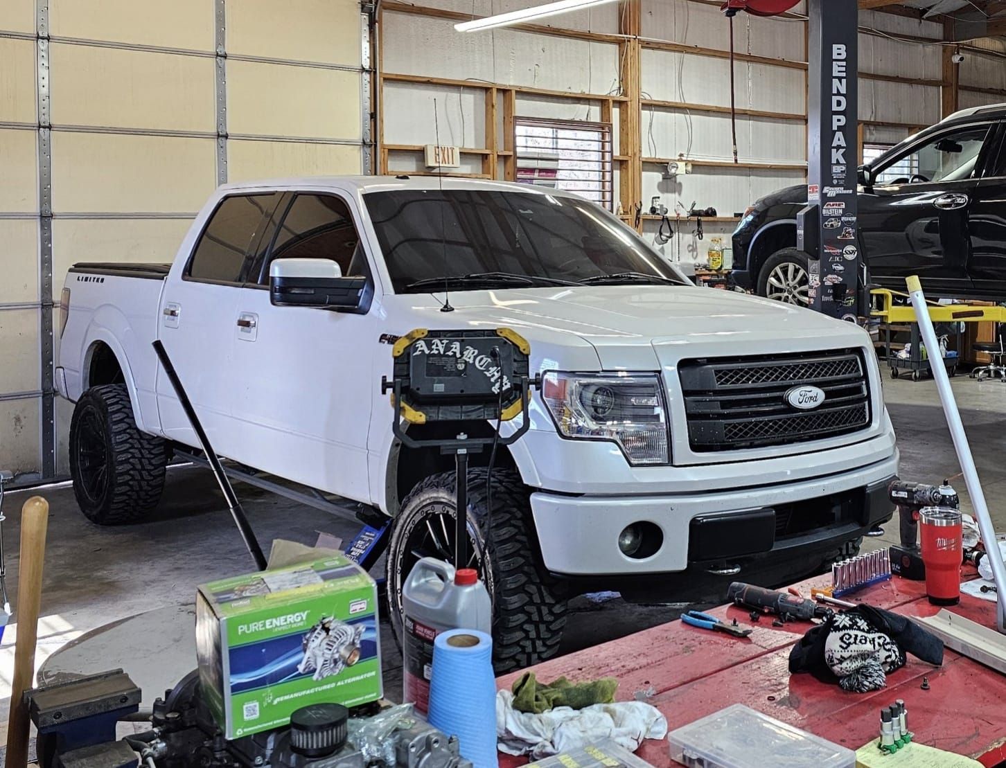 White Ford F-150 in a garage; front end lifted, tools on a red table, shop interior.