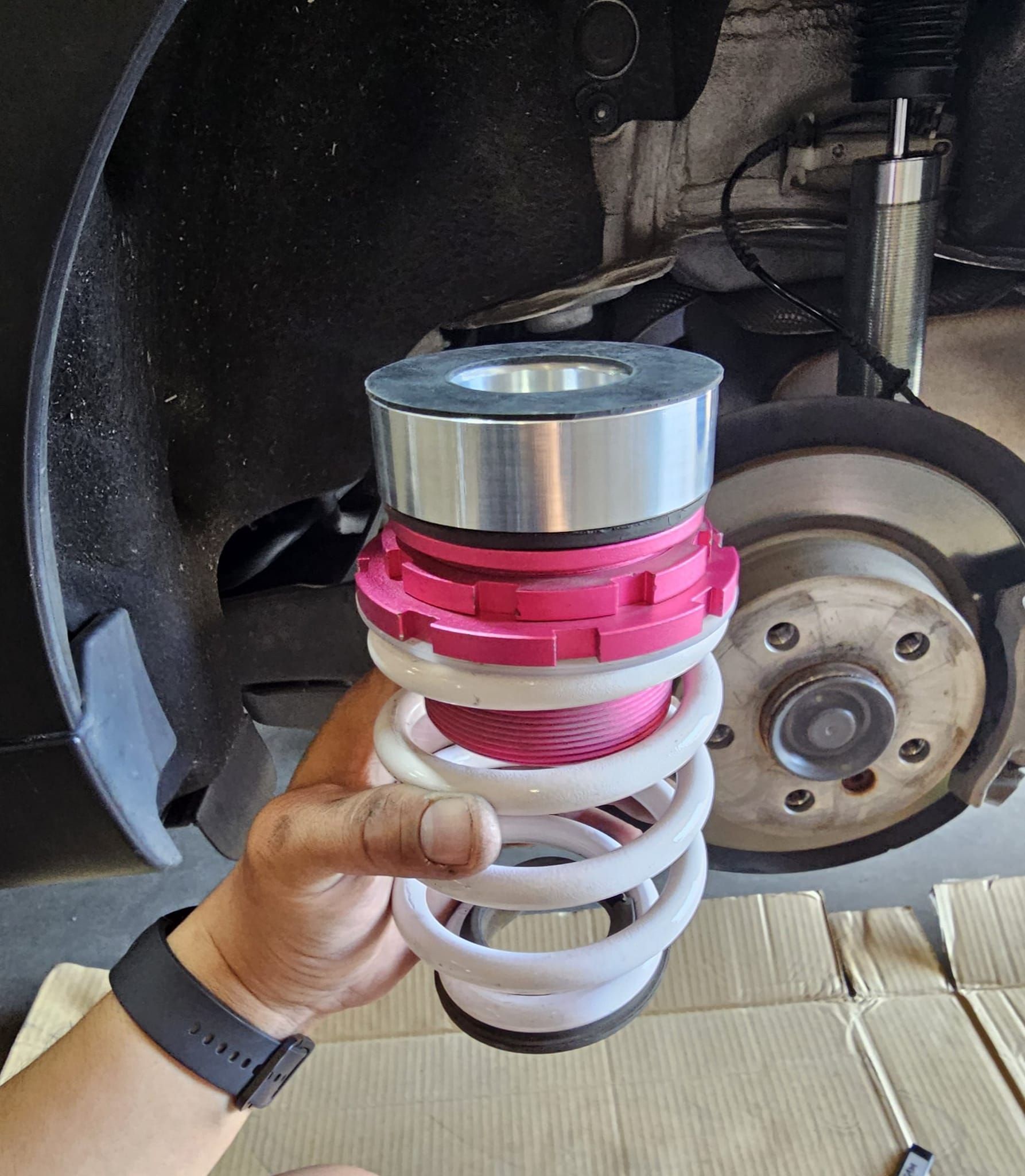 A hand holding a white and pink coilover suspension system with a car wheel and tire in the background.