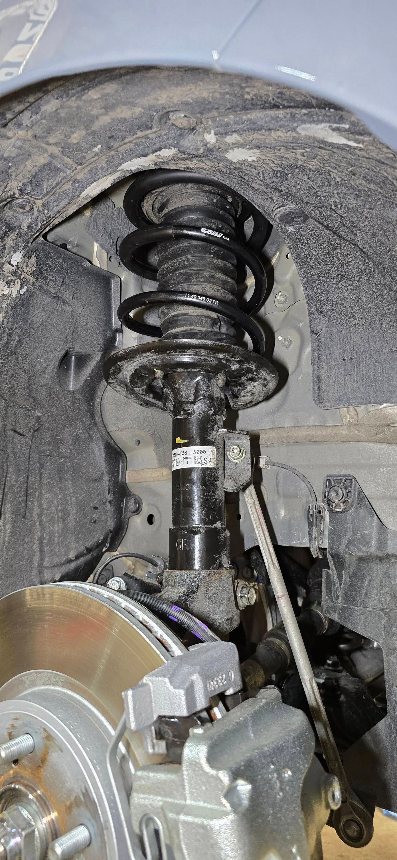 Car suspension with shock absorber, coil spring, brake disc, and caliper visible.