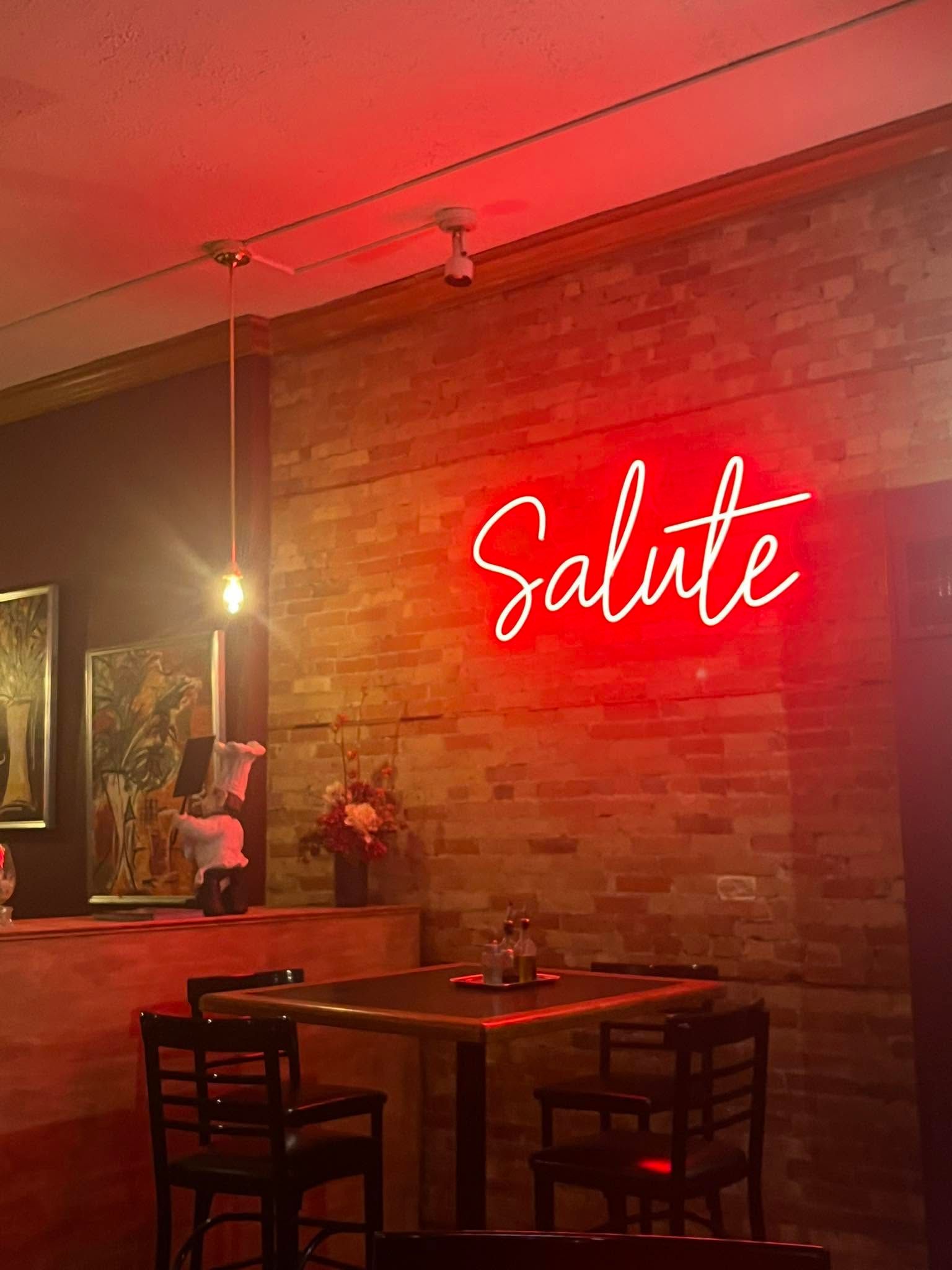 Red neon “Salute” sign above a small table and chairs in a warm brick-walled restaurant.
