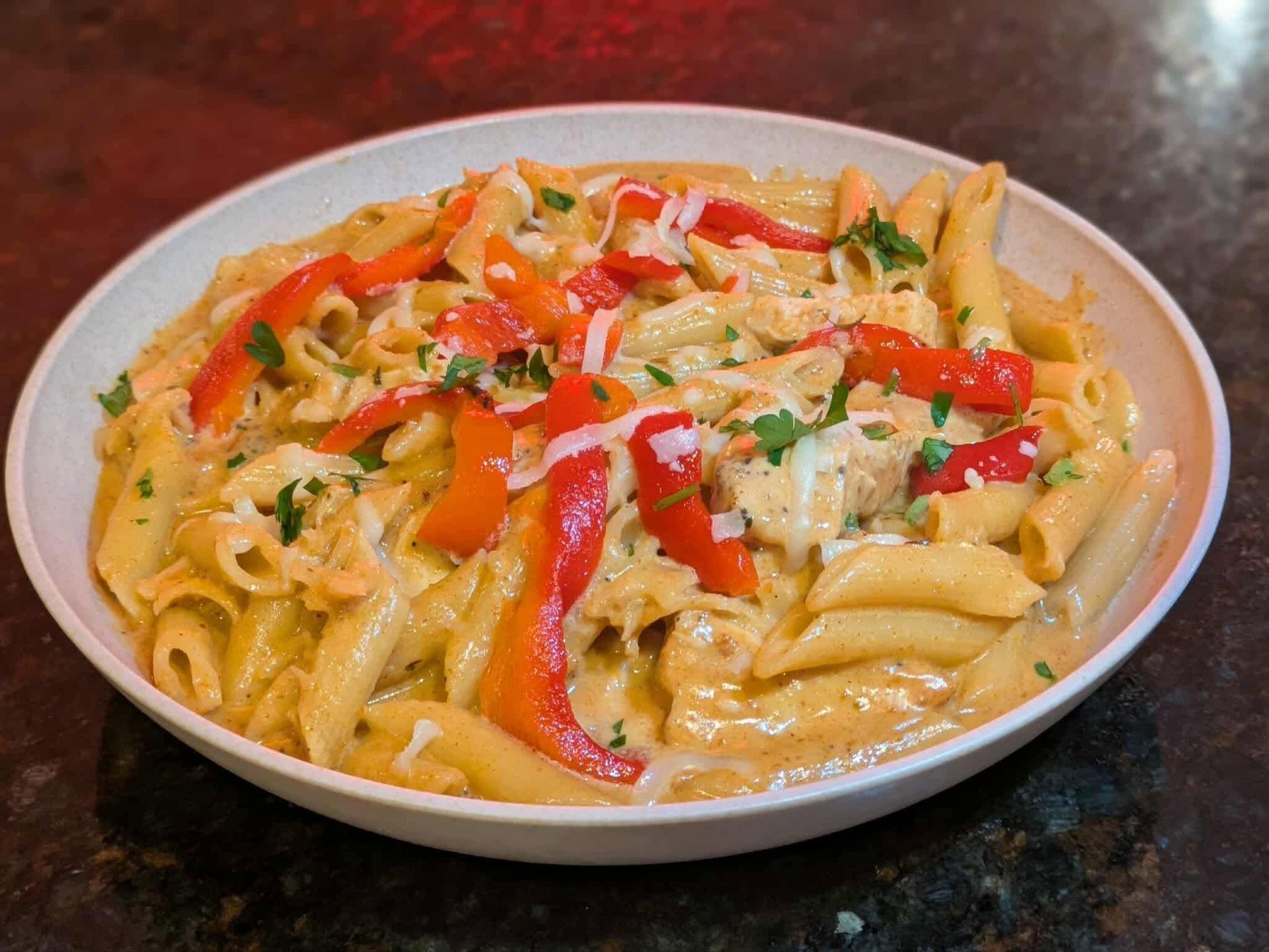 Plate of penne pasta with creamy sauce, red peppers, and herbs on a dark tabletop