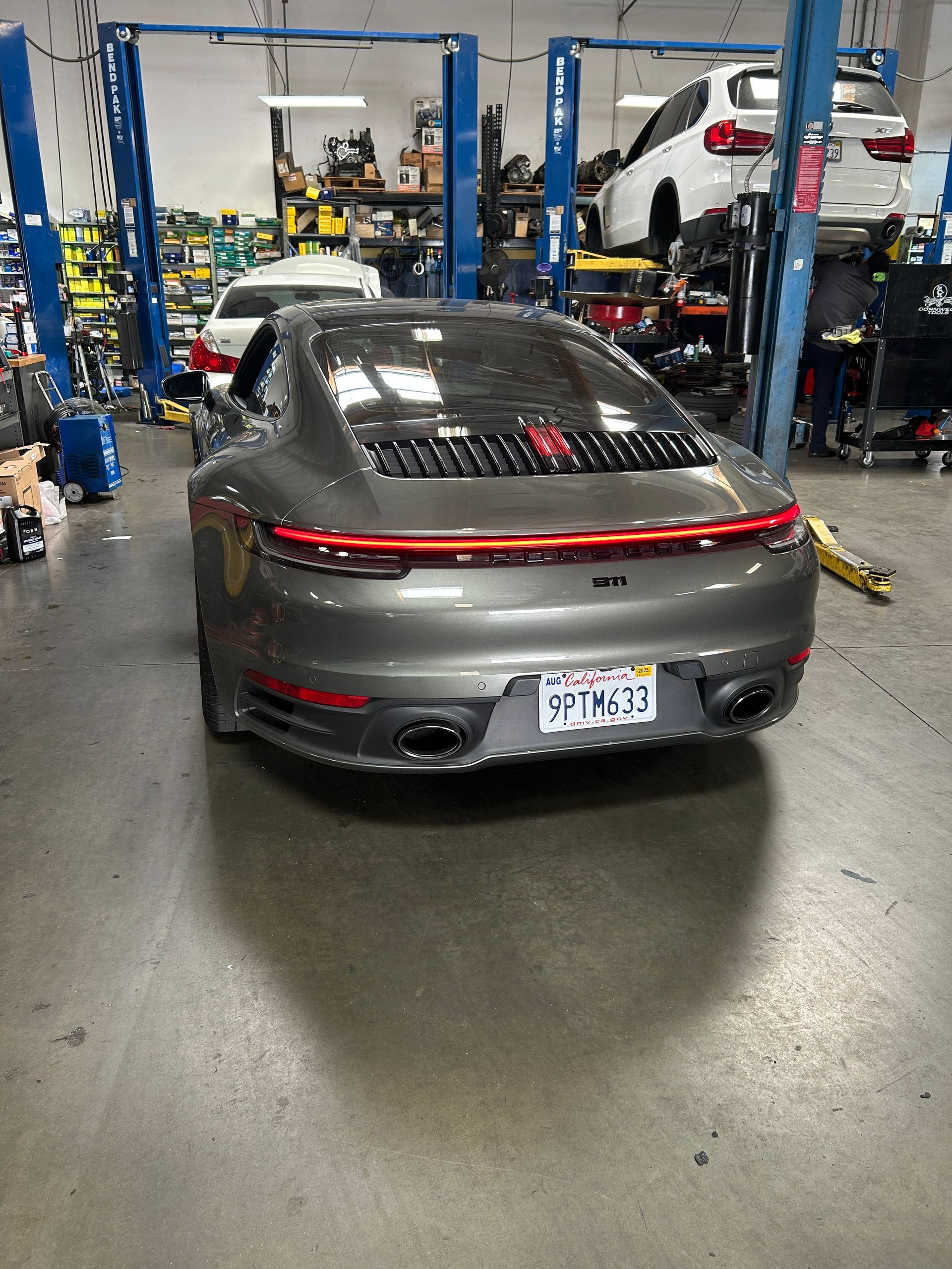 Gray sports car in a workshop bay, rear view with red taillight strip and lifted SUV nearby.