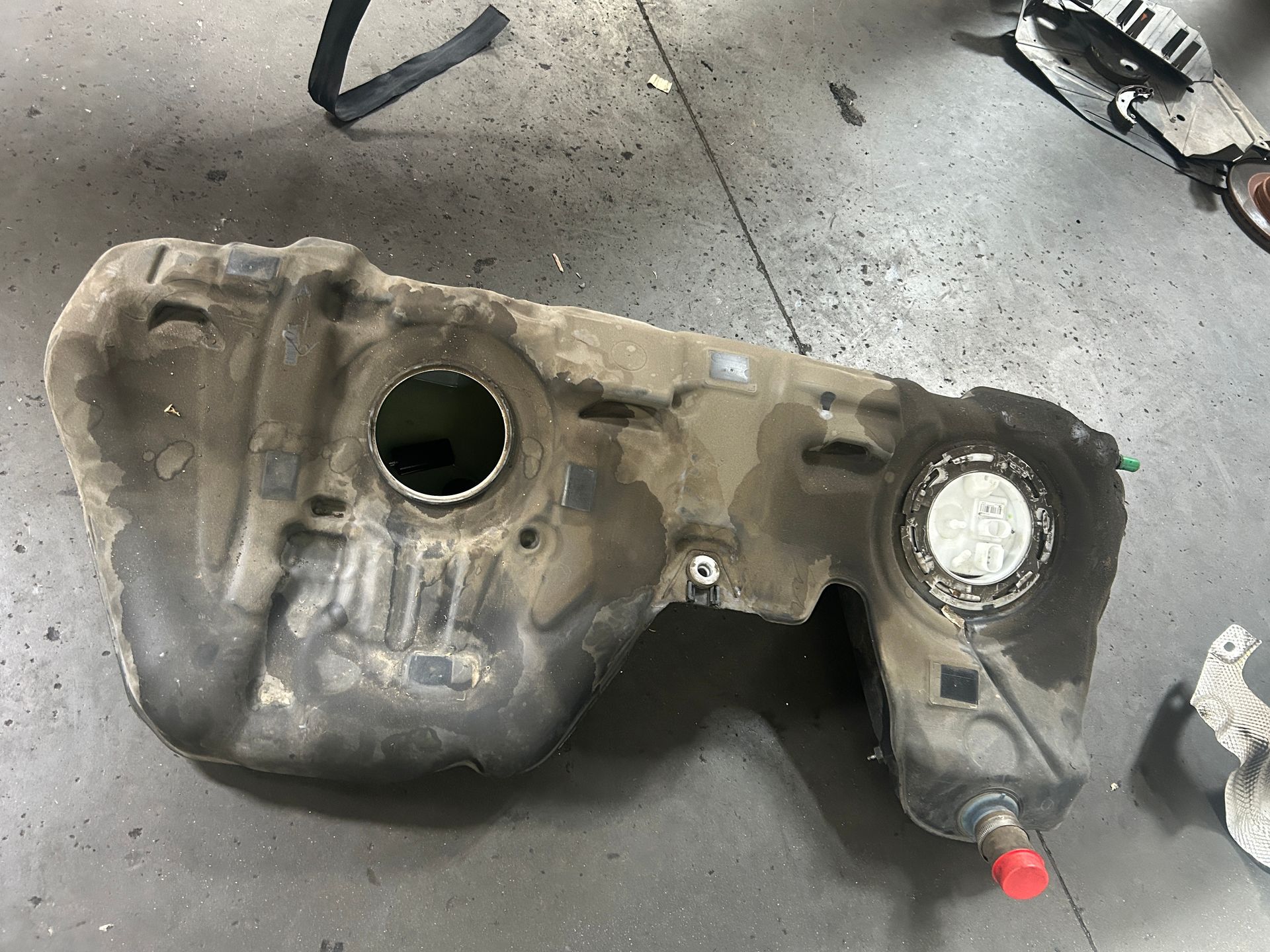 Dirty fuel tank on a garage floor with two circular openings and a red fitting.