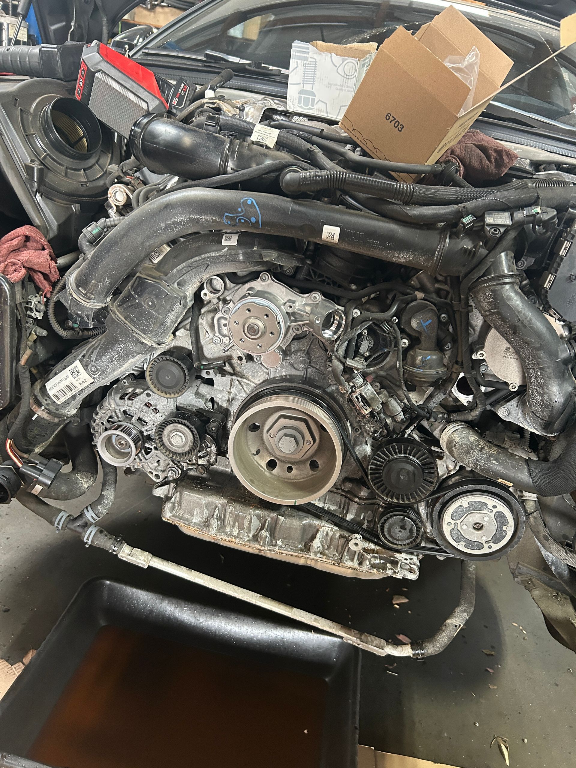 Exposed car engine front with serpentine belt and hoses, in a garage with a cardboard box on top.