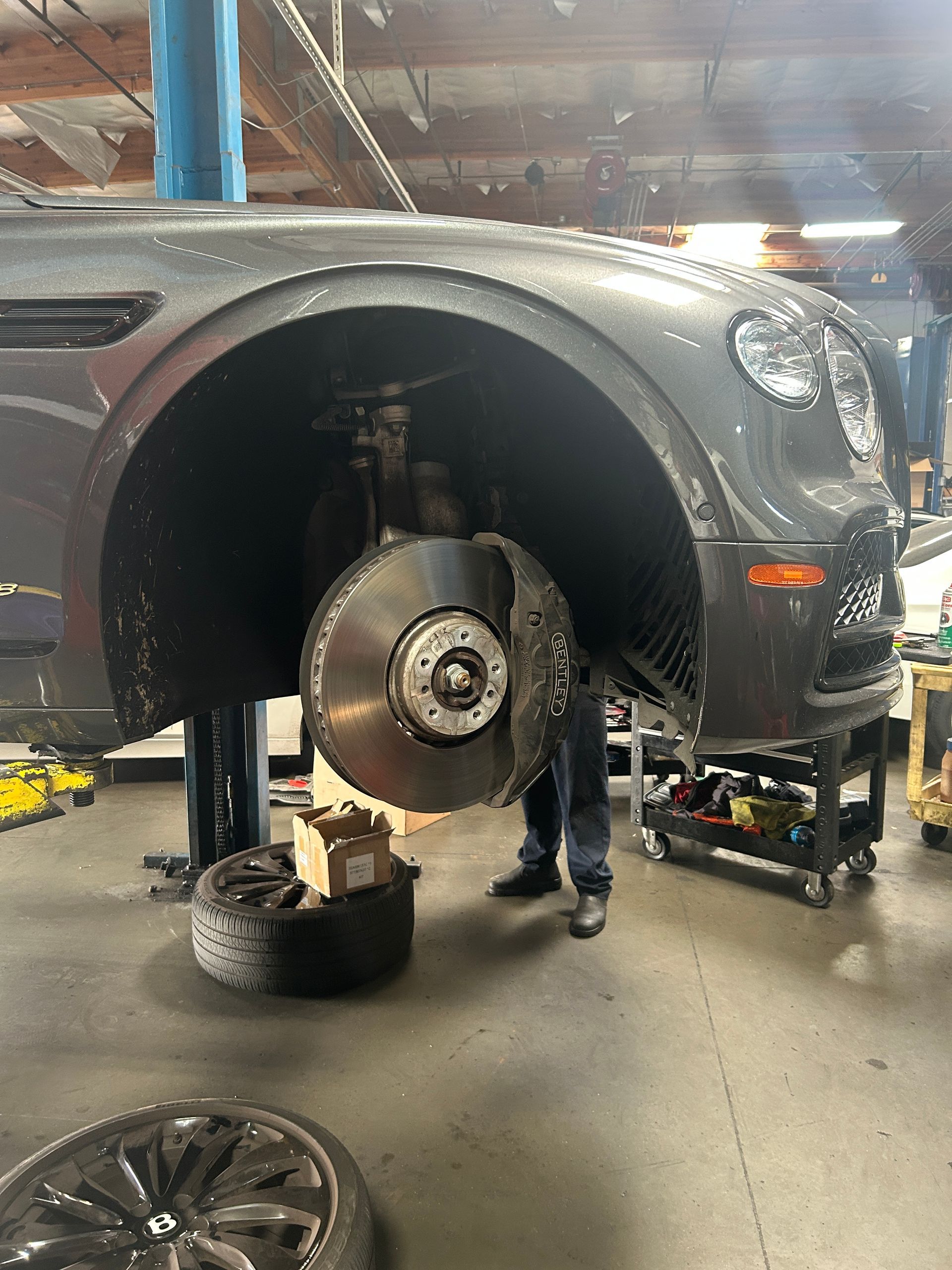 Car in auto shop lifted with front wheel removed; mechanic works underneath.
