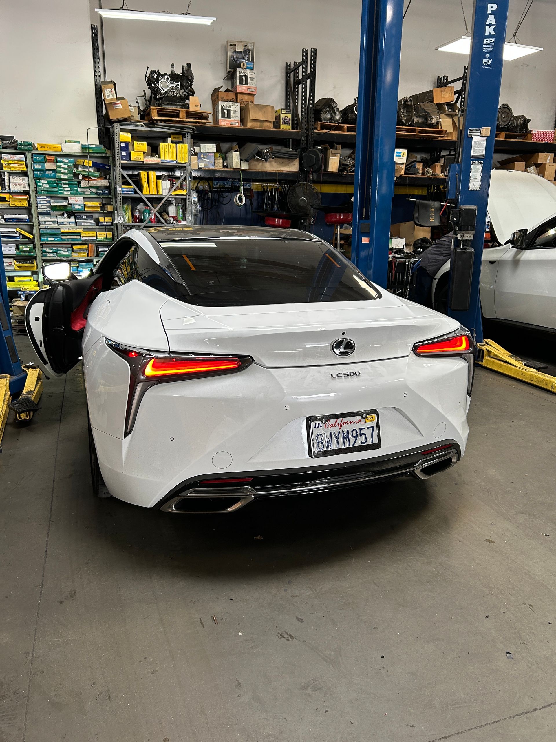 White sedan in a garage workshop, viewed from the rear with shelves and tools in the background