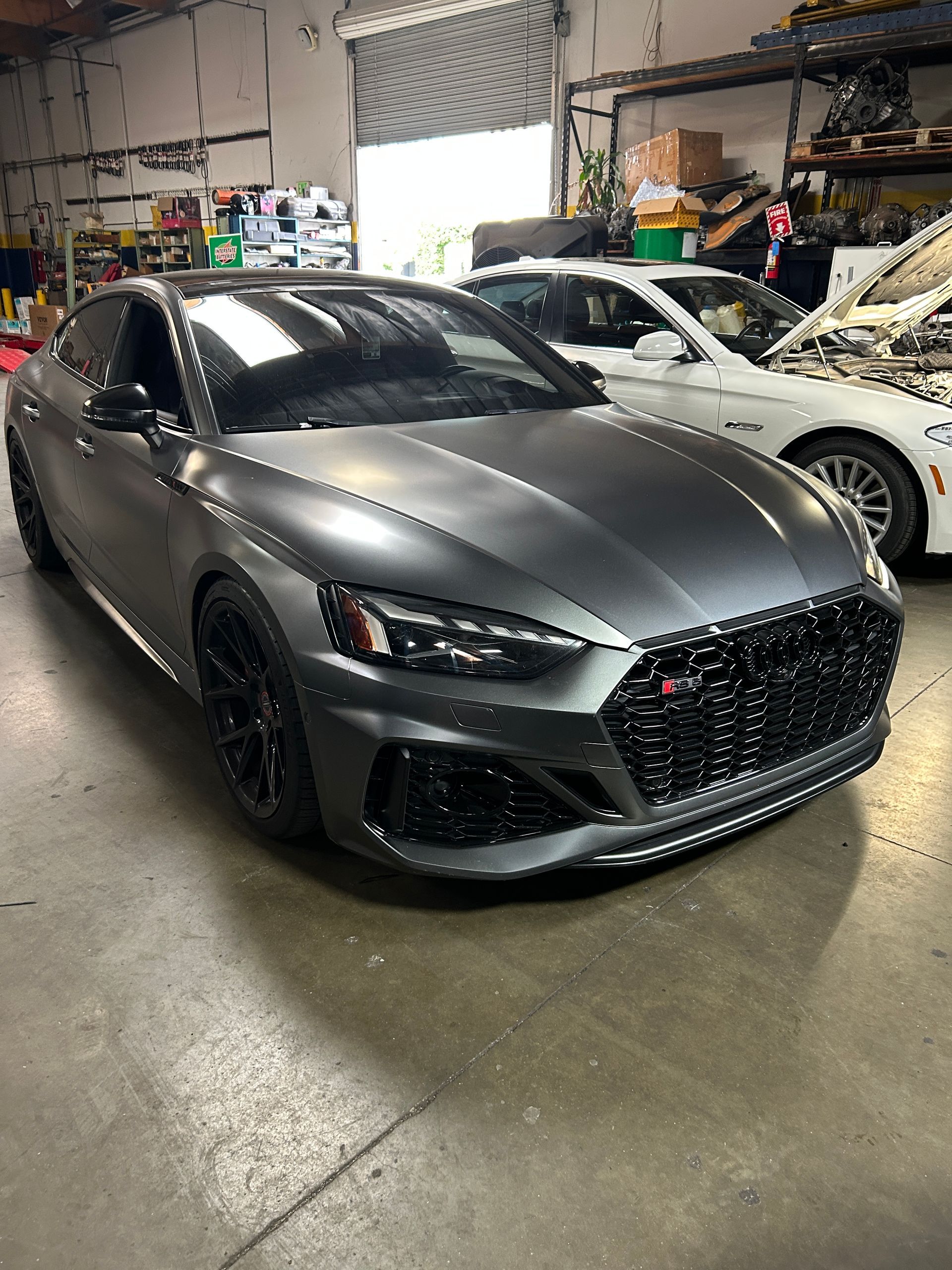 Gray sports sedan in a garage, with a white car parked beside it.