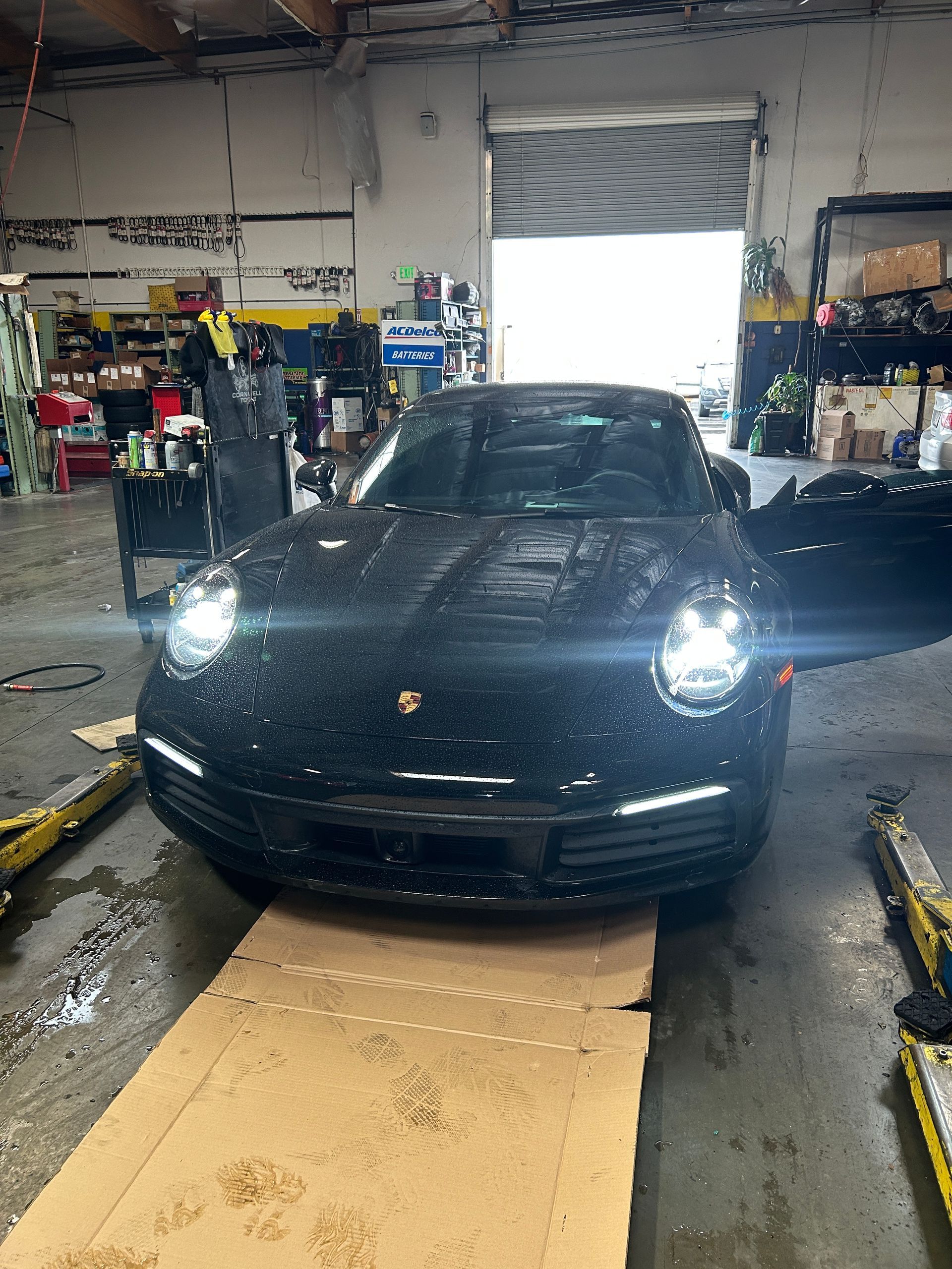 Black Porsche in a garage with headlights on, parked on a cardboard-covered floor and one door open