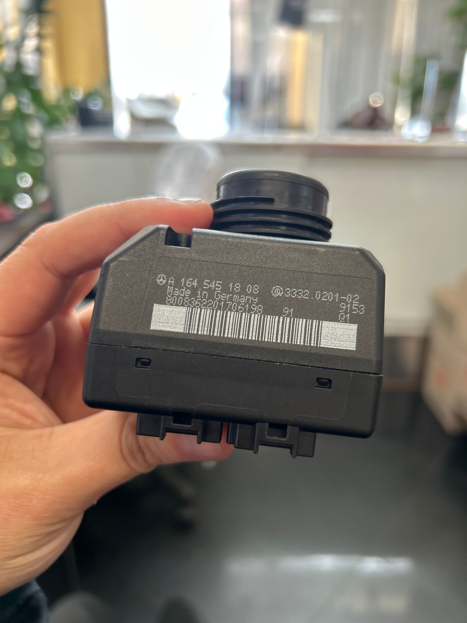 Hand holding a small black electronic module with connectors and a barcode label, indoors.