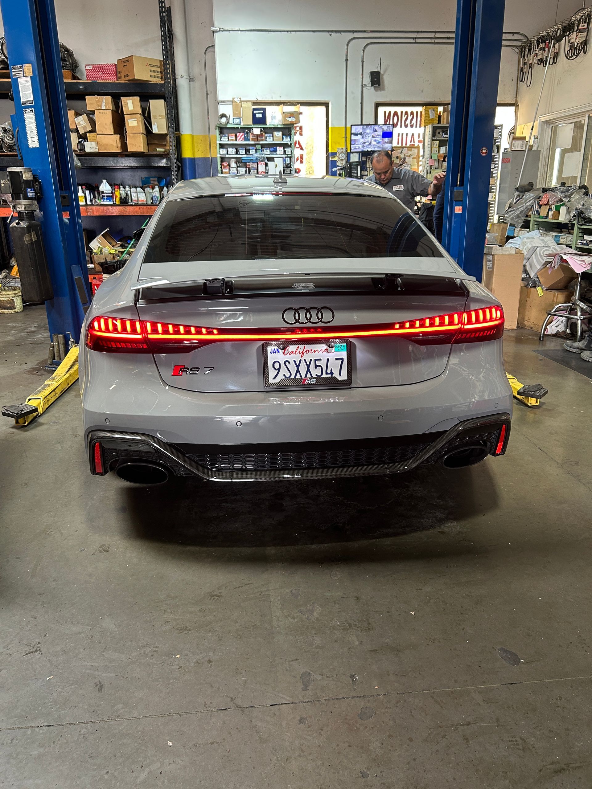 Gray sedan parked in a garage, rear view with illuminated taillights and shop equipment around it